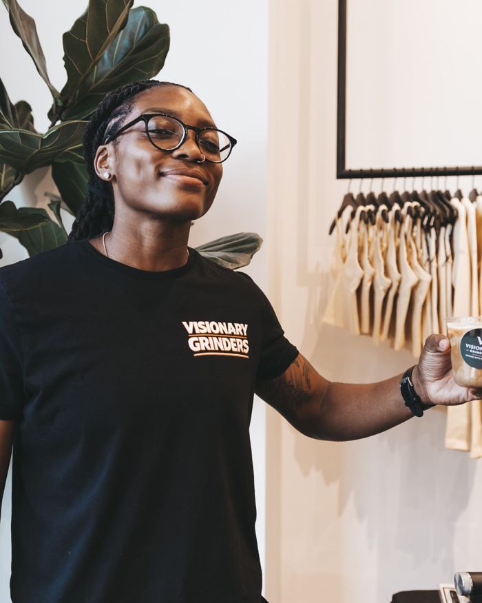 Jasmine Rhodes in a Visionary Grinders shirt next to a plant and clothing rack